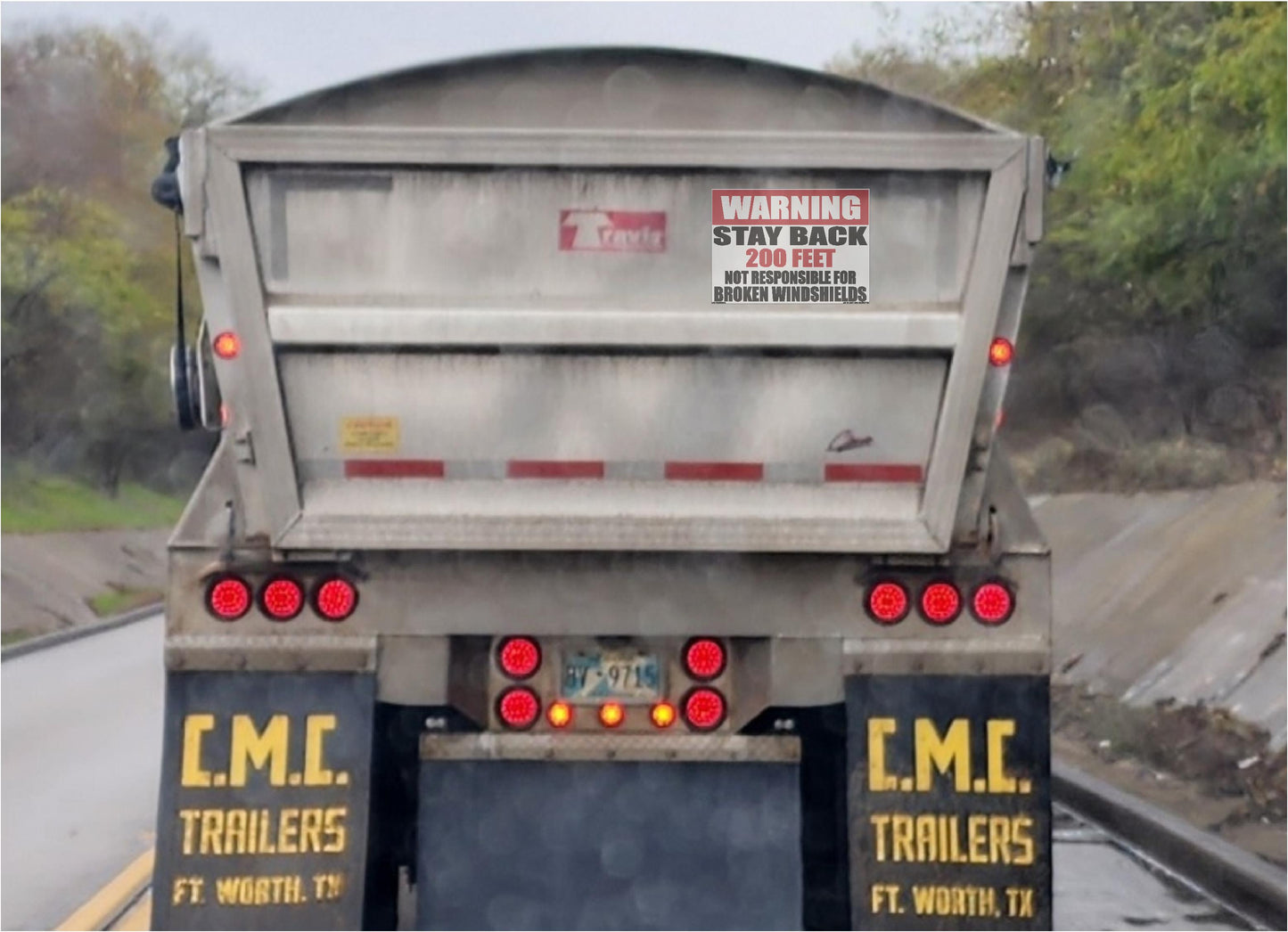 Rear view of a large dump truck on a road with trees in the background. The truck has a gray metal dump bed with a large Warning Stay Back safety decal, red taillights, and black rear panels with yellow 'C.